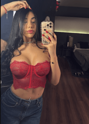 Woman with long dark hair wearing a red lace corset top and jeans, taking a mirror selfie in a bedroom.