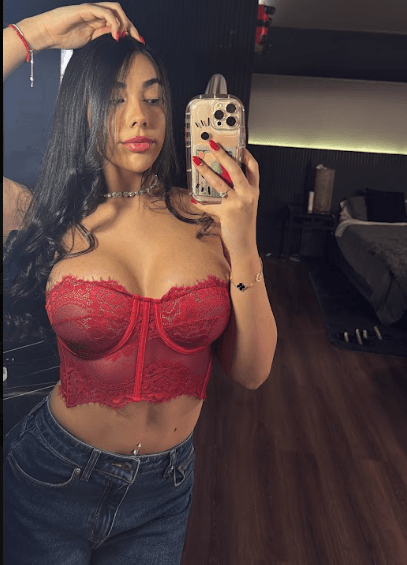 Woman with long dark hair wearing a red lace corset top and jeans, taking a mirror selfie in a bedroom.