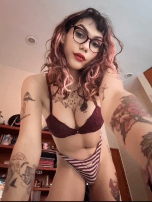 Person with pink-dyed hair and glasses poses in a burgundy lace bra and striped panties in a bedroom, showing tattoos on arms and chest.