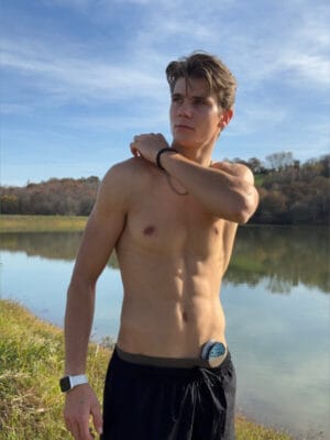 Young shirtless man by a lake, looking to the side, adjusting his shoulder, wearing a white smartwatch and black shorts; a small can sits at his waistband.