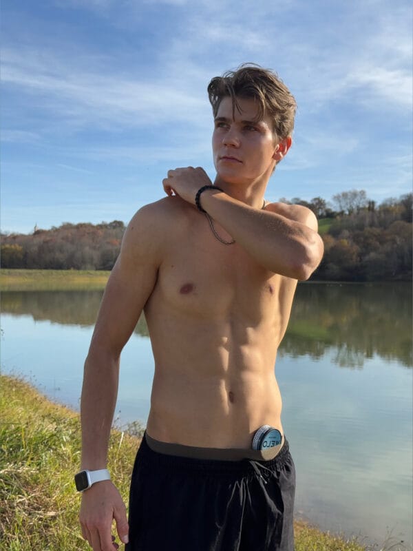 Young shirtless man by a lake, looking to the side, adjusting his shoulder, wearing a white smartwatch and black shorts; a small can sits at his waistband.