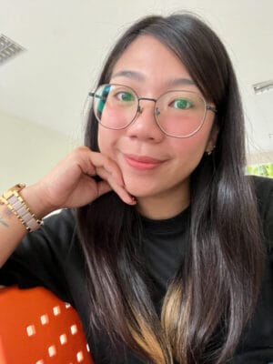 Woman with round glasses smiling, resting her chin on her hand, in a casual black shirt indoors.